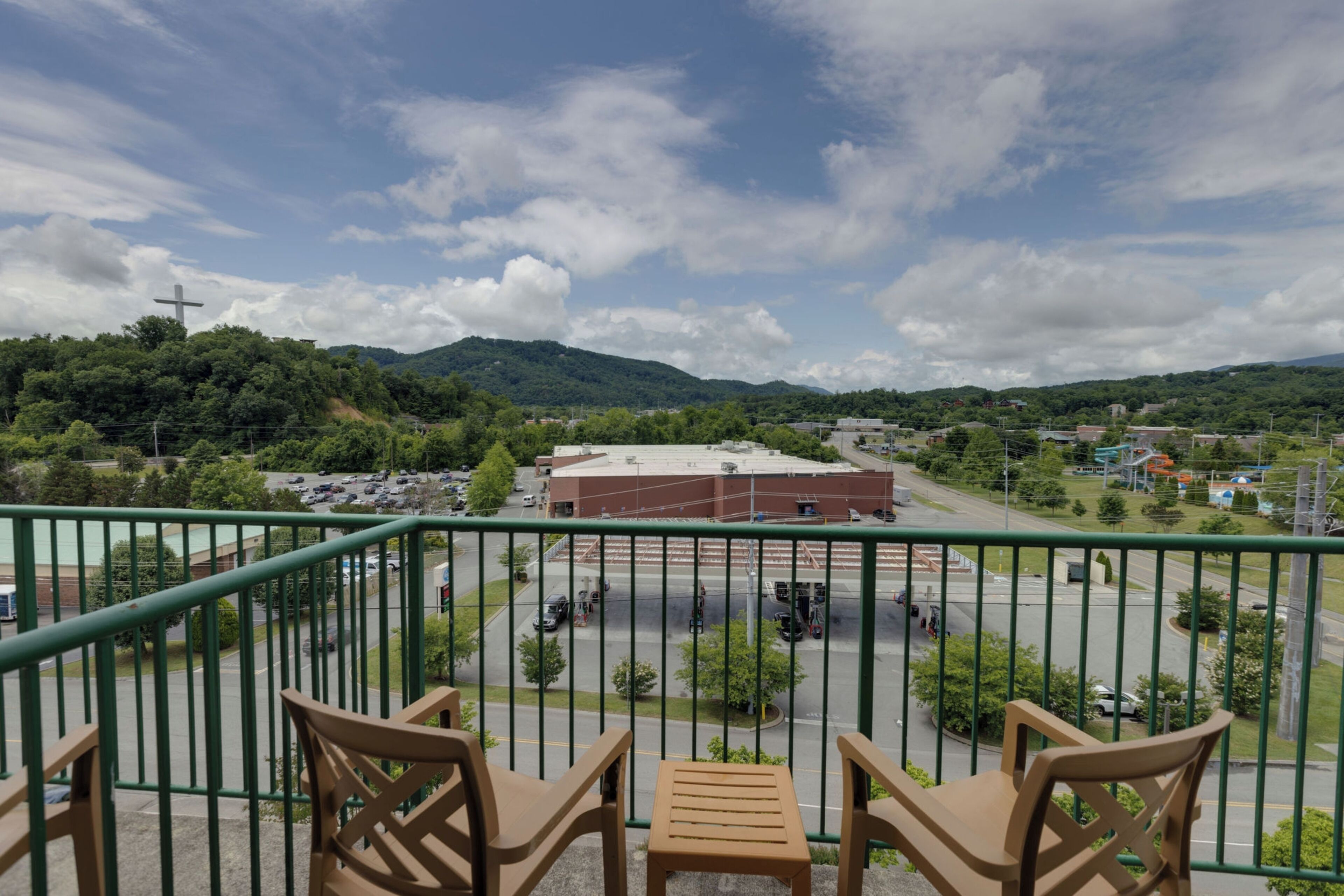 Un balcone con sedie di legno e vista su un parcheggio, un edificio e colline verdi sotto un cielo parzialmente nuvoloso.