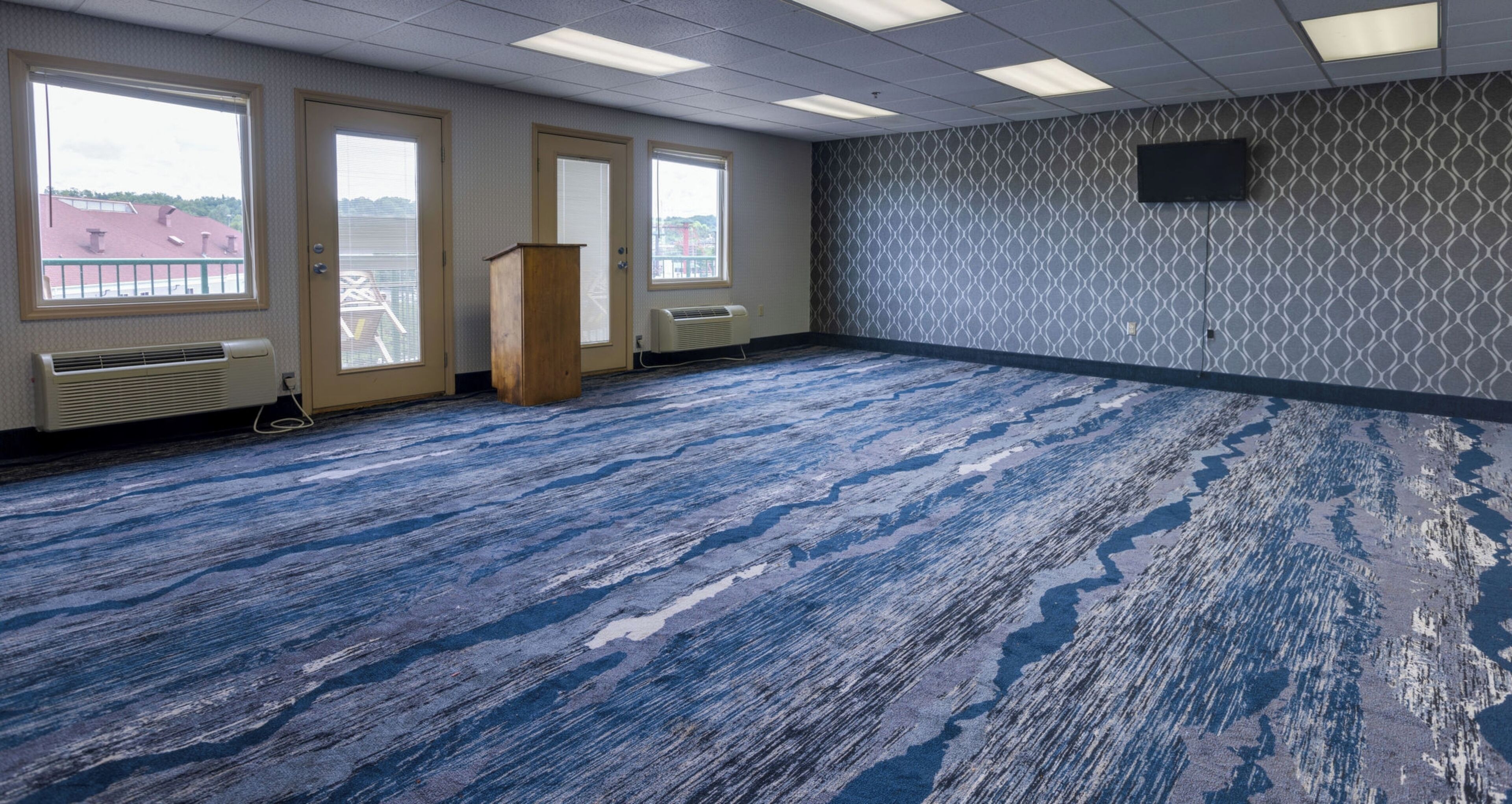 Un salón de conferencias con alfombra estampada, ventanas amplias y una televisión montada.
