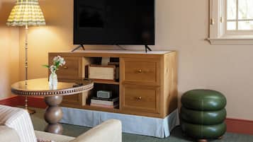 Un salon avec un téléviseur à écran plat, une table ronde en bois surmontée d’un vase, un pouf vert et une fenêtre ornée de rideaux à carreaux.