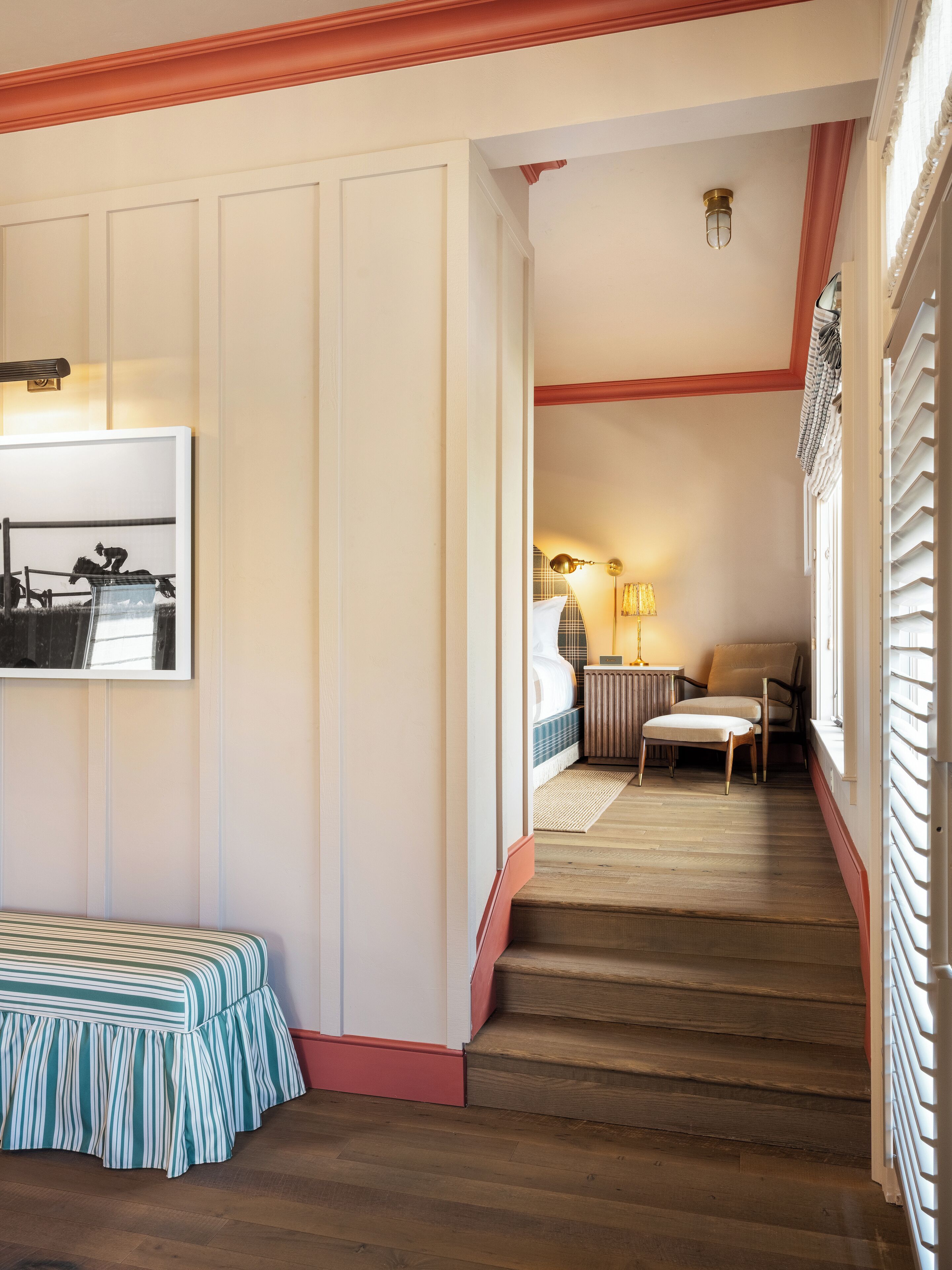 A hallway with a bench, a bed, and a chair in a room with wooden flooring and white walls.