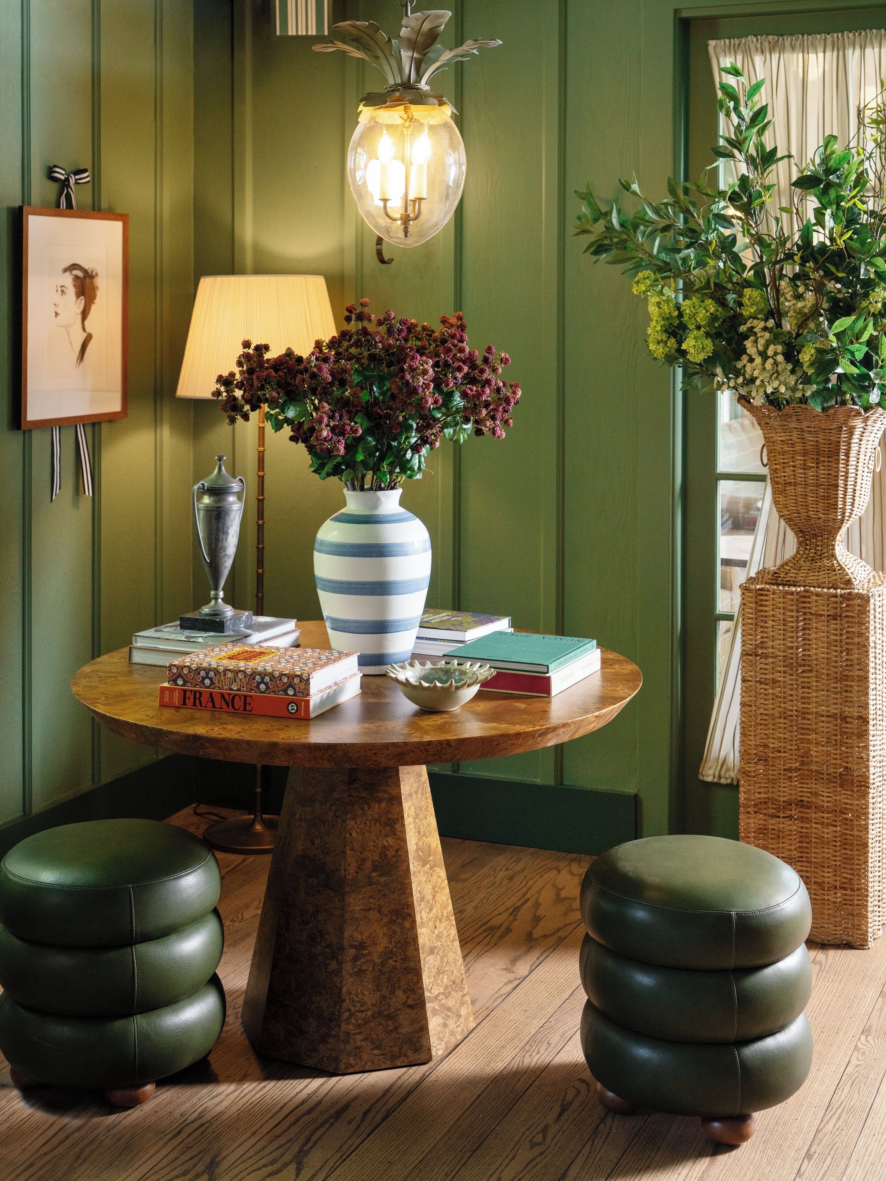 Una mesa redonda de madera con un jarrón de flores, libros y una lámpara.