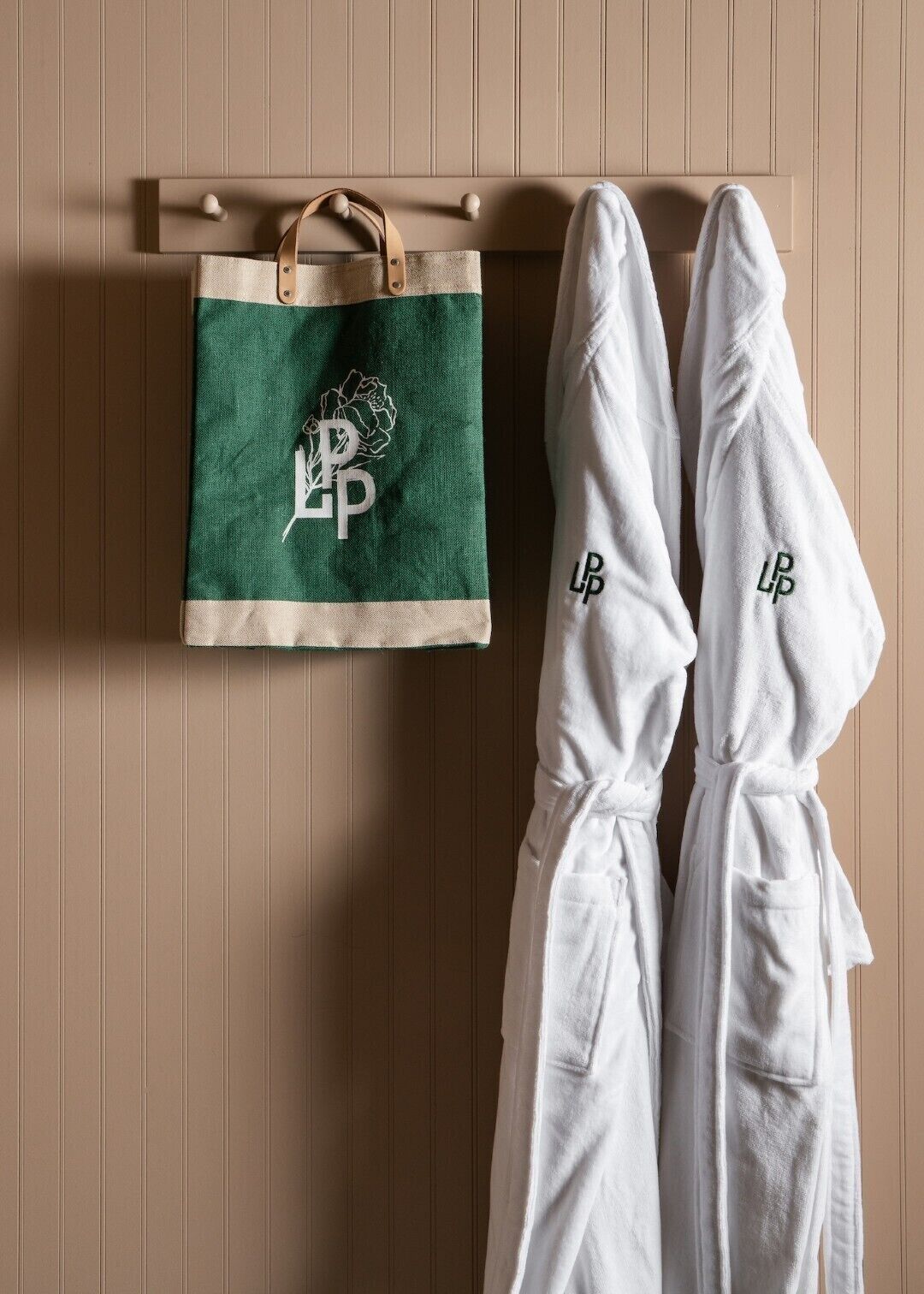 A bathroom with a wooden panel wall, a green and beige toiletry bag, and two white bathrobes with embroidered initials.