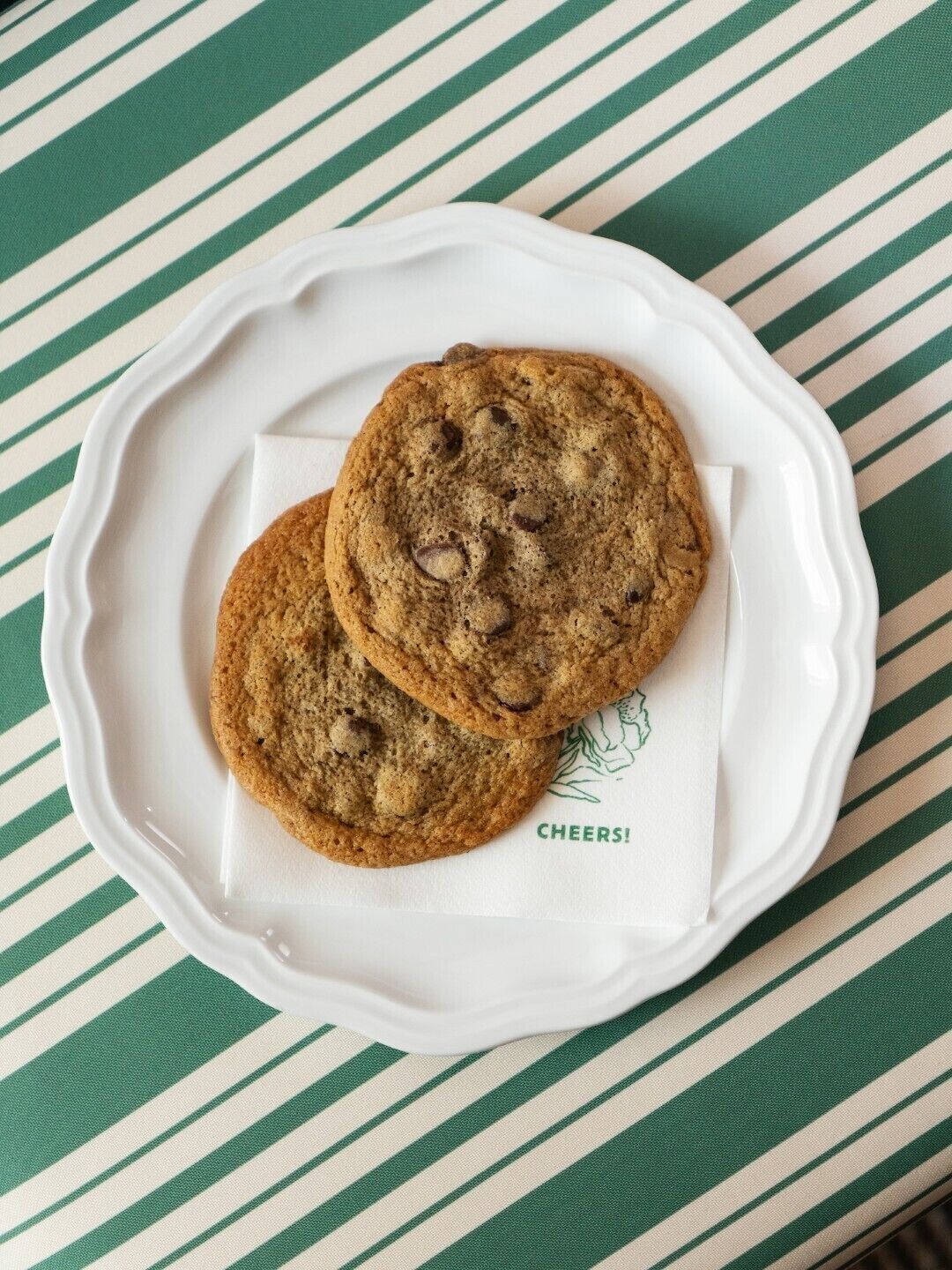 Two chocolate chip cookies on a white plate with a green and white striped tablecloth.
