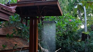 A wooden deck with a traditional roof structure, surrounded by lush greenery and a water feature. - Sugar Hut 1984 (Pattaya)