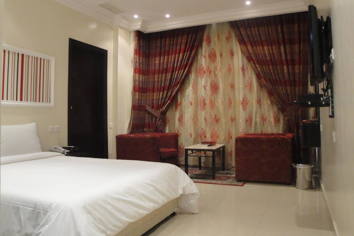 Ein Hotelzimmer mit einem Bett, zwei Sesseln, einem kleinen Tisch und einem an der Wand montierten Fernseher.
