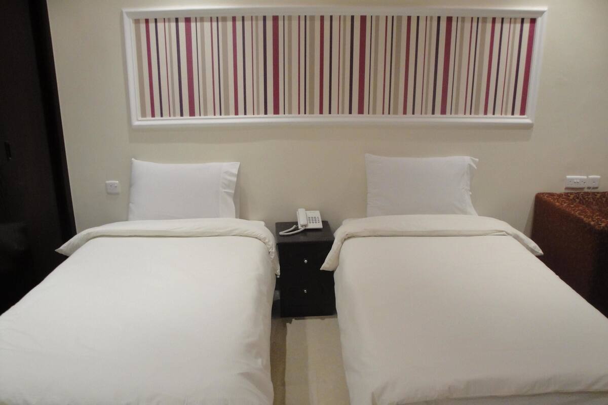 Ein Hotelzimmer mit zwei Einzelbetten, einem kleinen Nachttisch mit Telefon und einem Fenster mit gestreiften Jalousien.