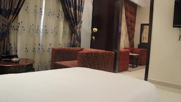 A hotel room with a bed, a small table, two armchairs, and a door leading to a sitting area with a red sofa.