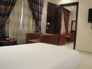 Ein Hotelzimmer mit einem Bett, einem kleinen Tisch, zwei Sesseln und einer Tür zu einem Sitzbereich mit einem roten Sofa.