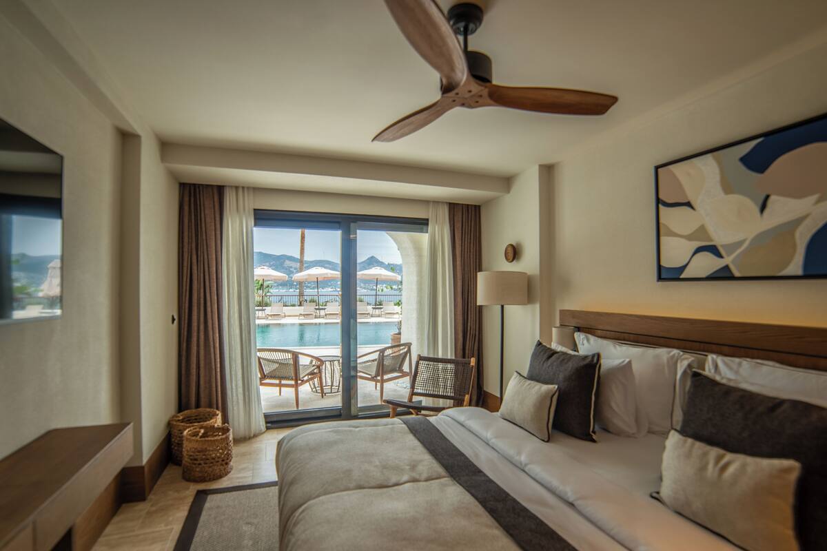 Quarto de hotel com cama, ventoinha no teto, vista para piscina e montanhas, e um quadro na parede.