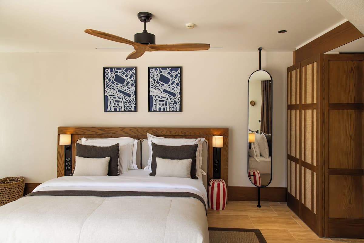 Quarto de hotel com cabeceira de madeira, cama com roupa de cama branca, ventilador de teto, duas obras de arte nas paredes, espelho e guarda-roupa de madeira.