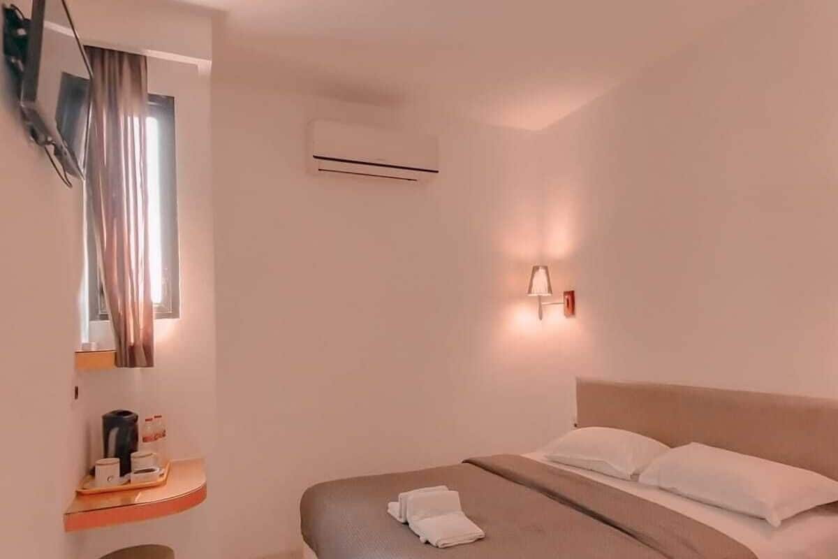 A modern hotel room with a bed, a small bedside table with a lamp, an air conditioning unit, and a television mounted on the wall.