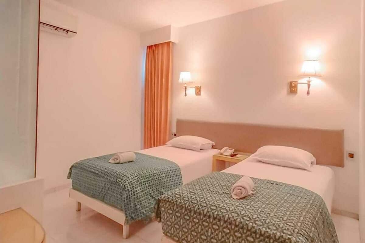 A hotel room with two beds, each with a patterned bedspread and a rolled towel. There are two wall-mounted lamps and a window with curtains.