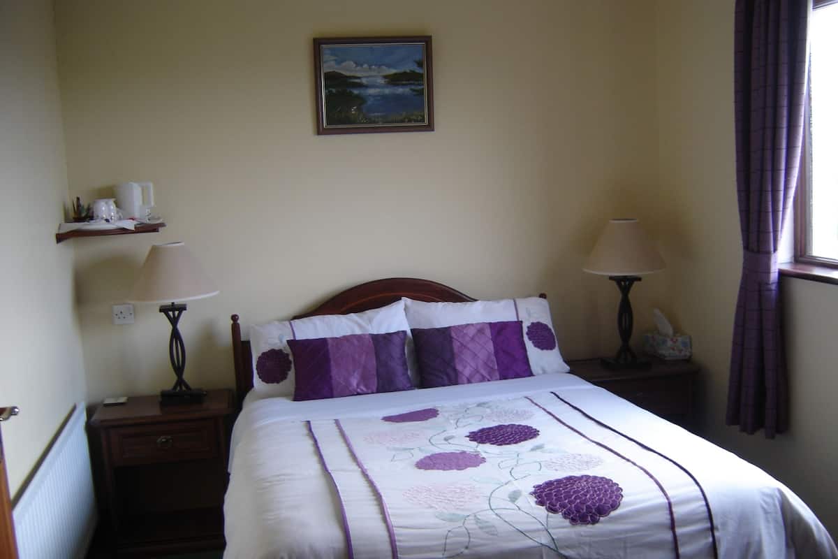 A neatly made bed with a floral bedspread, two bedside lamps, a nightstand, a shelf with items, and a painting on the wall.