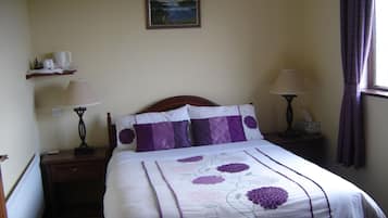 A neatly made bed with a floral bedspread, two bedside lamps, a nightstand, a shelf with items, and a painting on the wall.