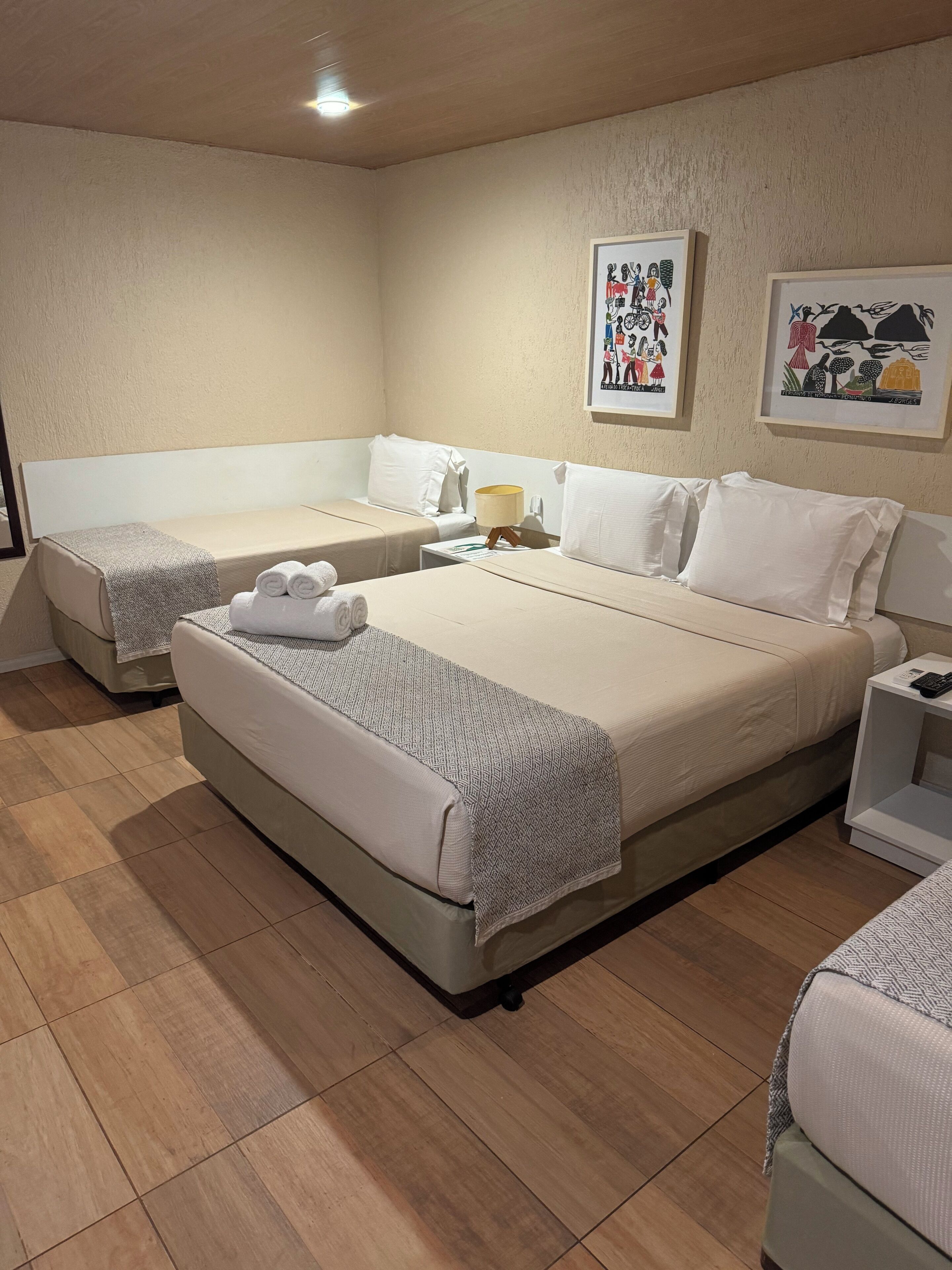 A hotel room with a large bed, a smaller bed, and a bedside table with a lamp. There are two framed pictures on the wall.