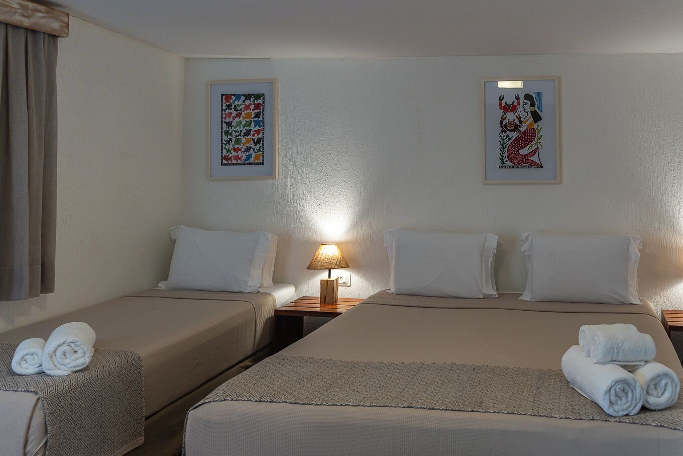 A hotel room with two single beds, a nightstand with a lamp, and two framed artworks on the walls.