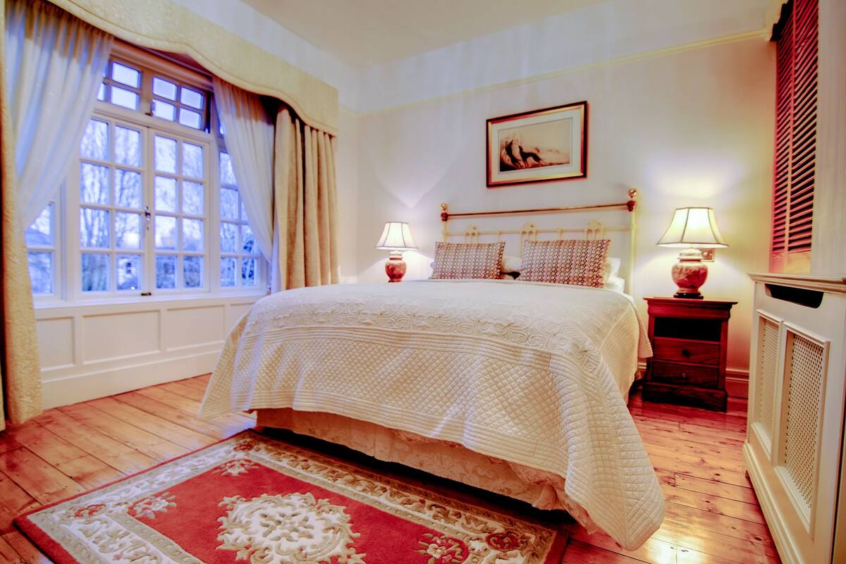 A bedroom with a large bed, two bedside tables with lamps, a window with curtains, a painting on the wall, and a radiator.