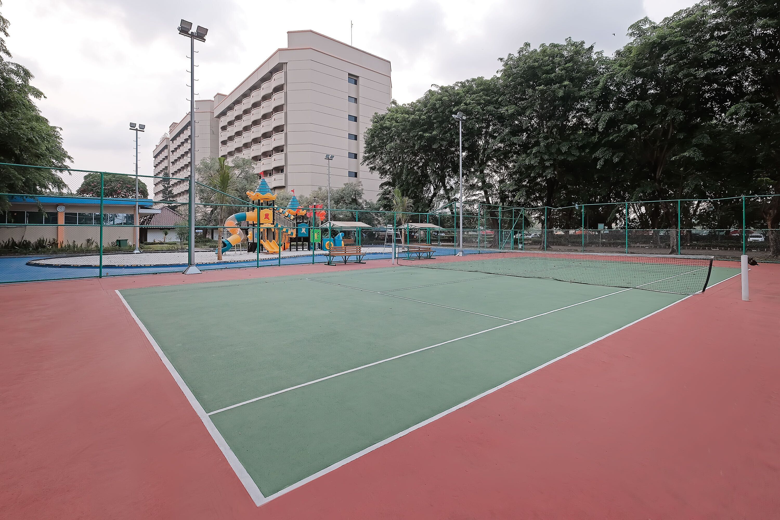 A tennis court with a net, surrounded by a fence, and a playground with slides and climbing structures.