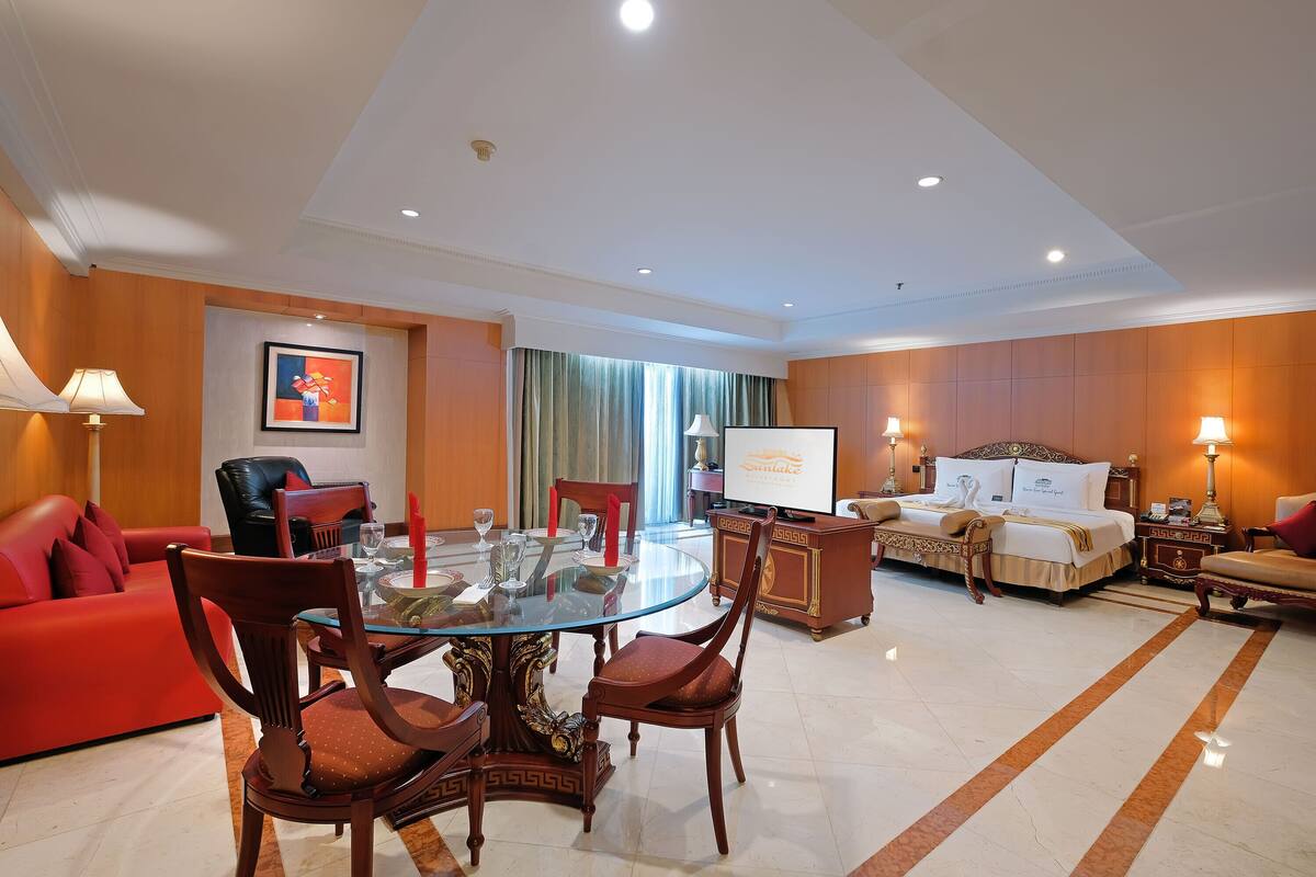 A hotel room with a glass dining table, red sofa, and a bed with a white comforter.