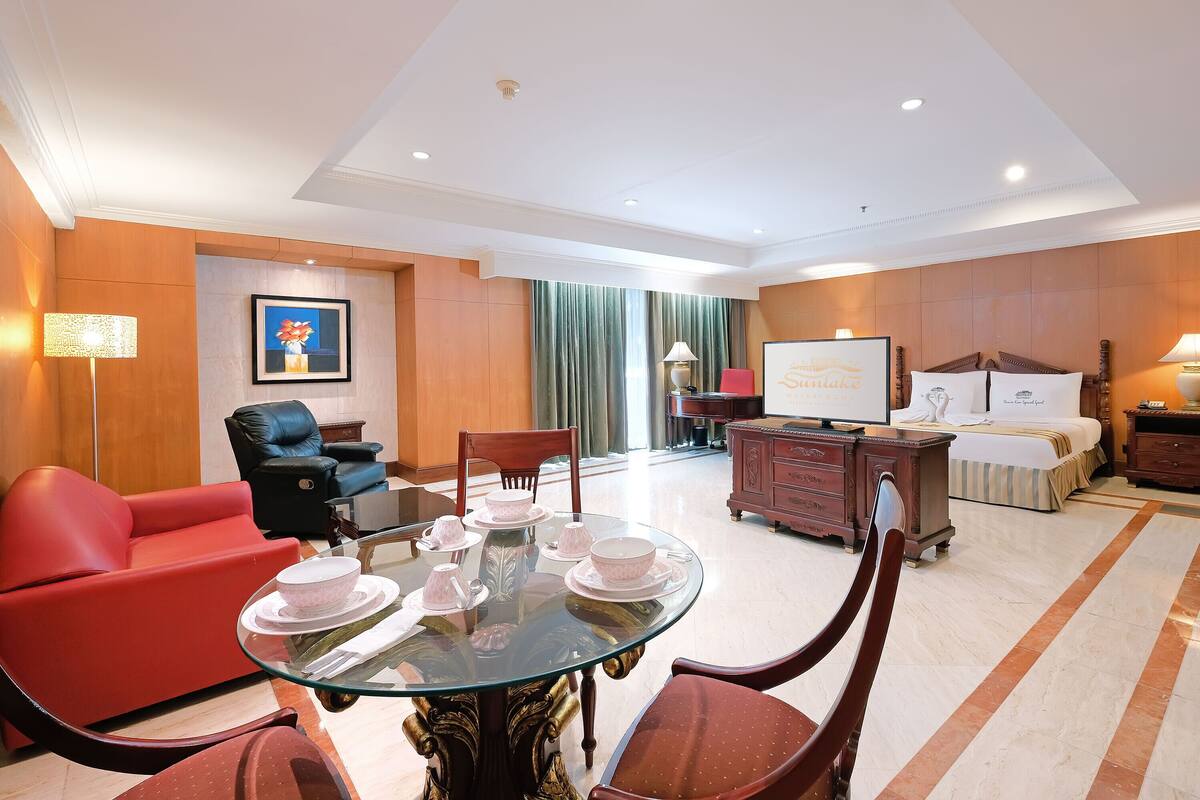 A hotel room with a glass-top dining table set with pink teacups and saucers, a red sofa, a black leather chair, a wooden cabinet, and a bed with a white comforter and pillows.