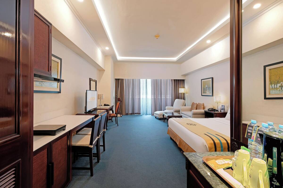 A hotel room with a desk, two chairs, a bed, a nightstand, a lamp, and a window with curtains.