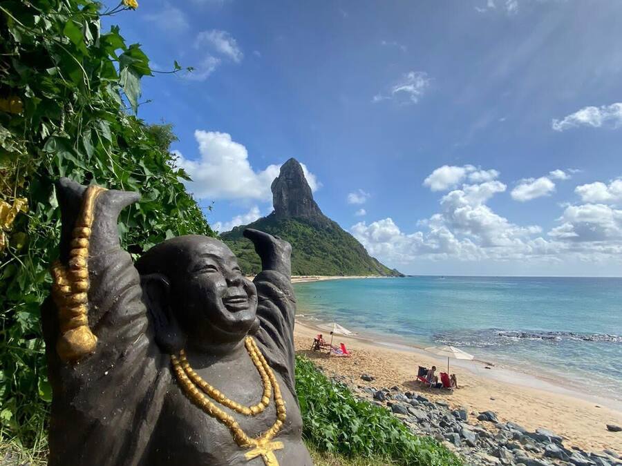 Una estatua de una figura con los brazos levantados, una cruz y un rosario, que domina una playa con aguas cristalinas y una montaña al fondo.