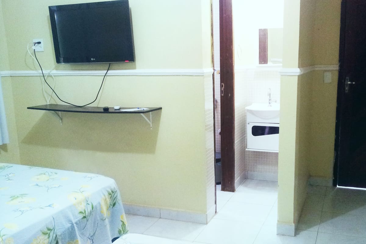 Une chambre d’hôtel avec un lit, un téléviseur fixé au mur, une petite table et une salle de bain visible à travers une porte ouverte.