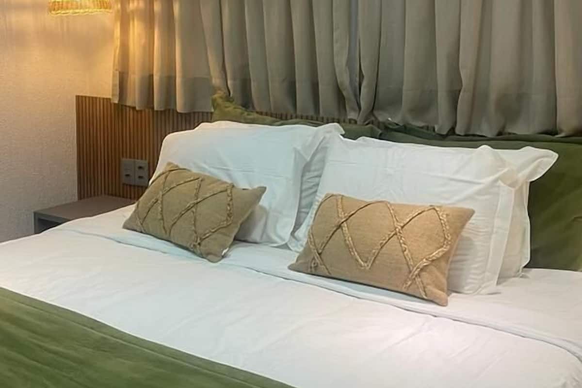Ein ordentlich bezogenes Bett mit weißer und grüner Bettwäsche, eine Hängelampe und ein großes Fenster mit Vorhängen.