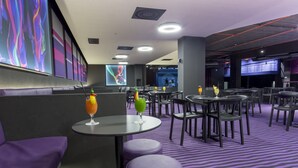 Ein modernes Restaurant mit violetten Sitzplätzen, Tischen mit Getränken und einem großen Bildschirm, der abstrakte Kunst zeigt.