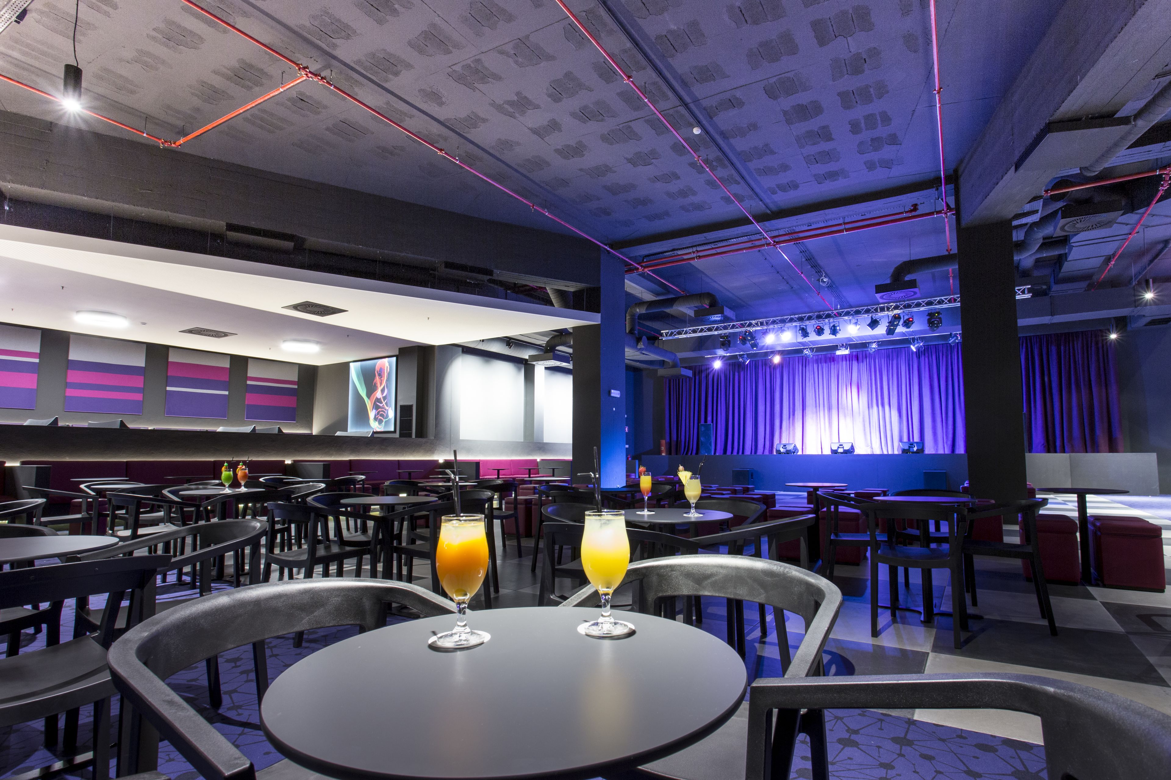 A modern restaurant with multiple tables and chairs, a stage with curtains, and a bar area.