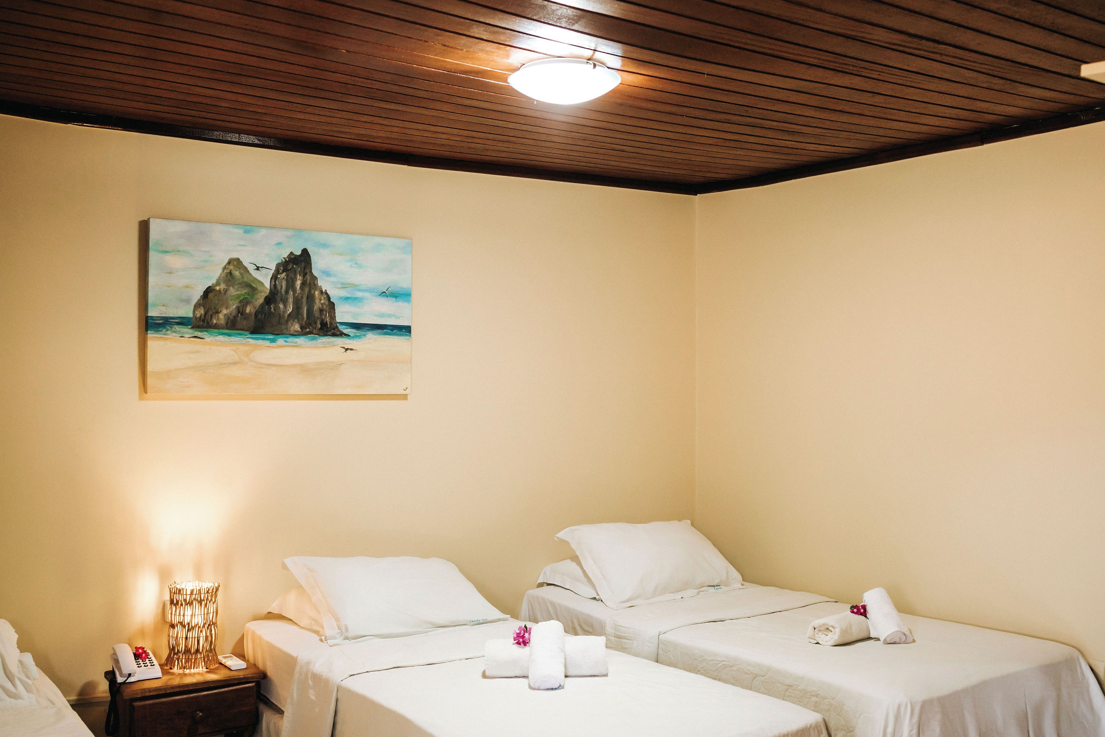A hotel room with two single beds, a nightstand with a lamp, a painting of a coastal scene, and a wooden ceiling.