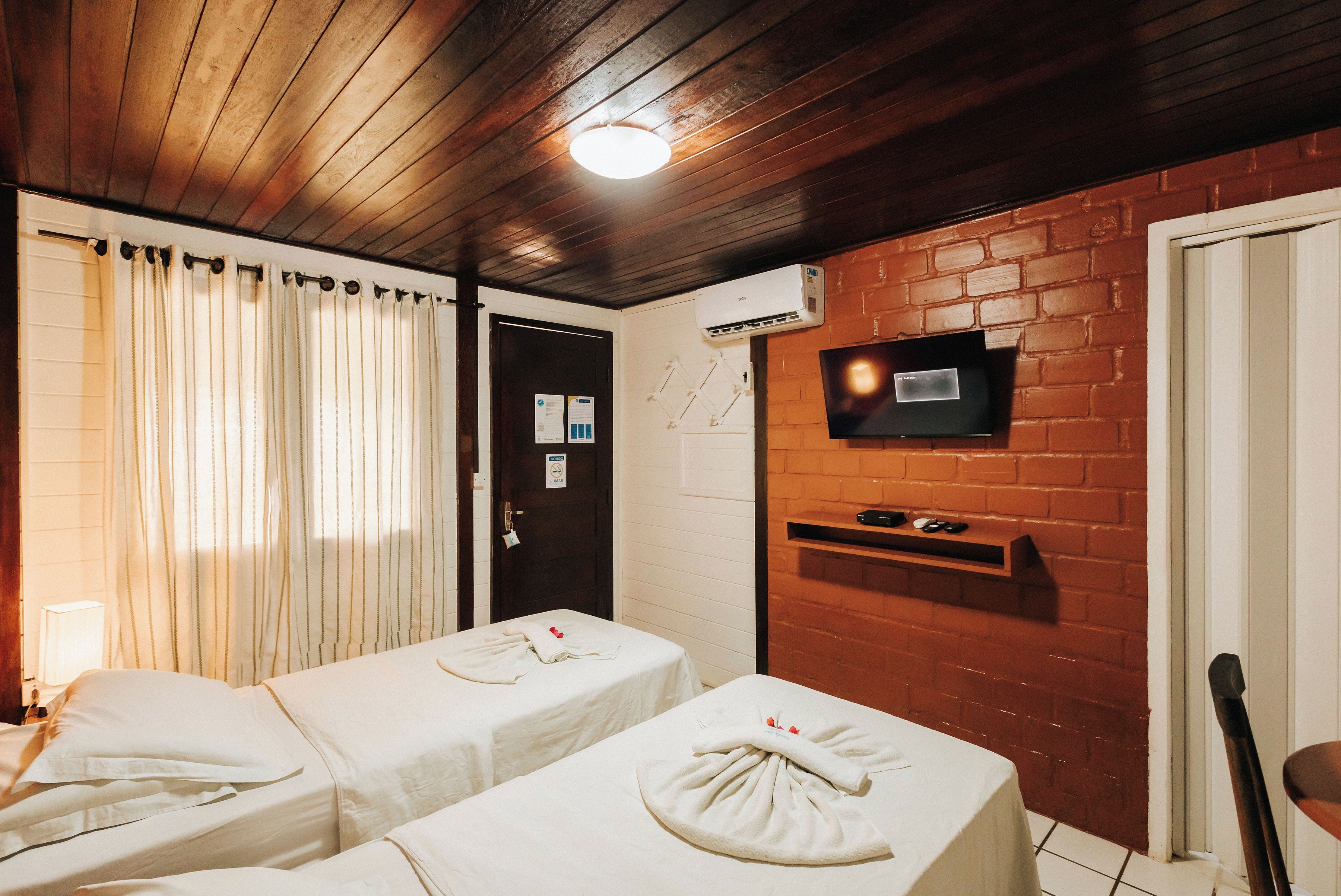 A hotel room with two beds, a television mounted on the brick wall, and an air conditioning unit.