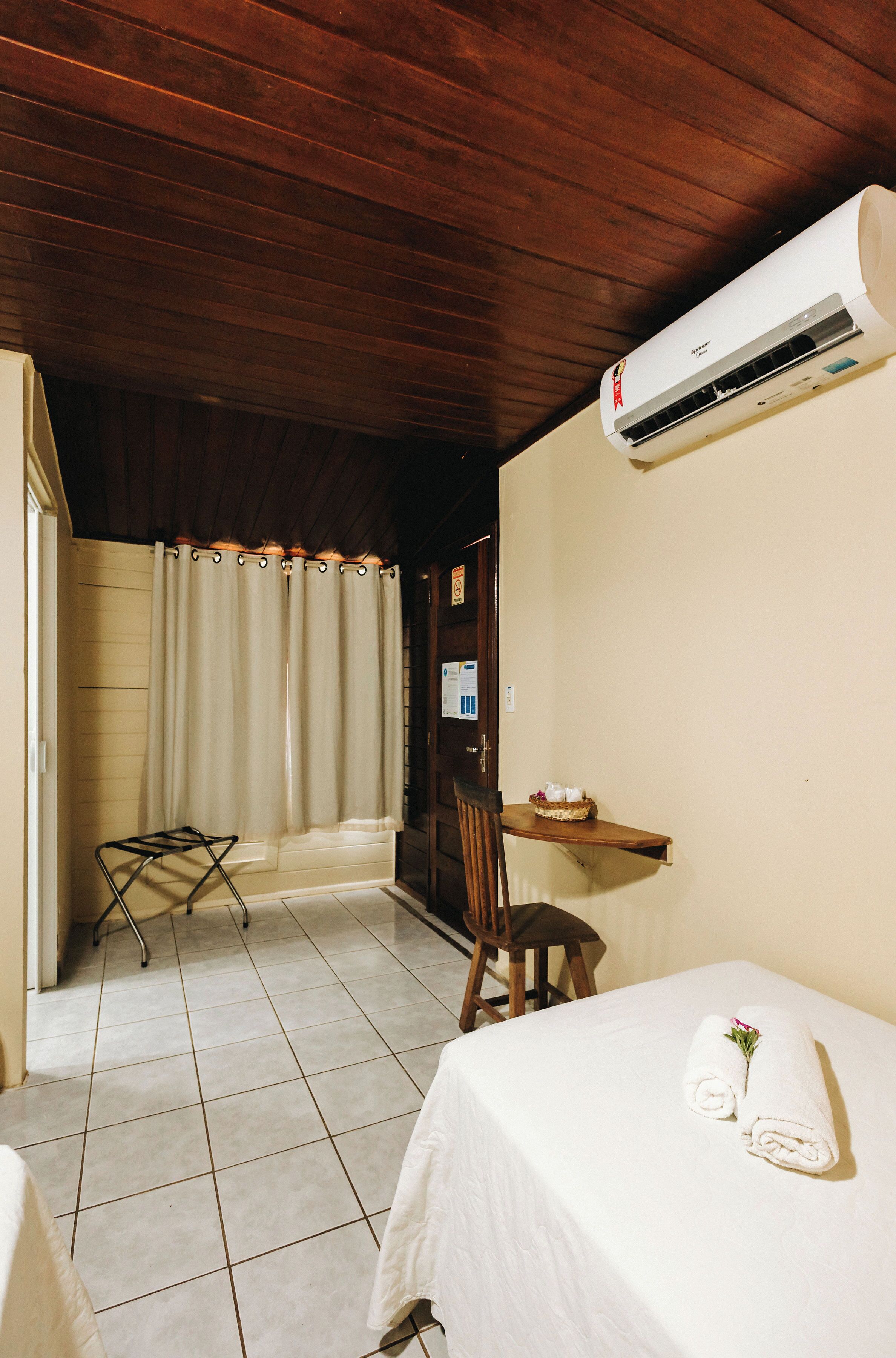 A hotel room with a wooden ceiling, a bed with white linens, a small table with a basket, and an air conditioning unit.