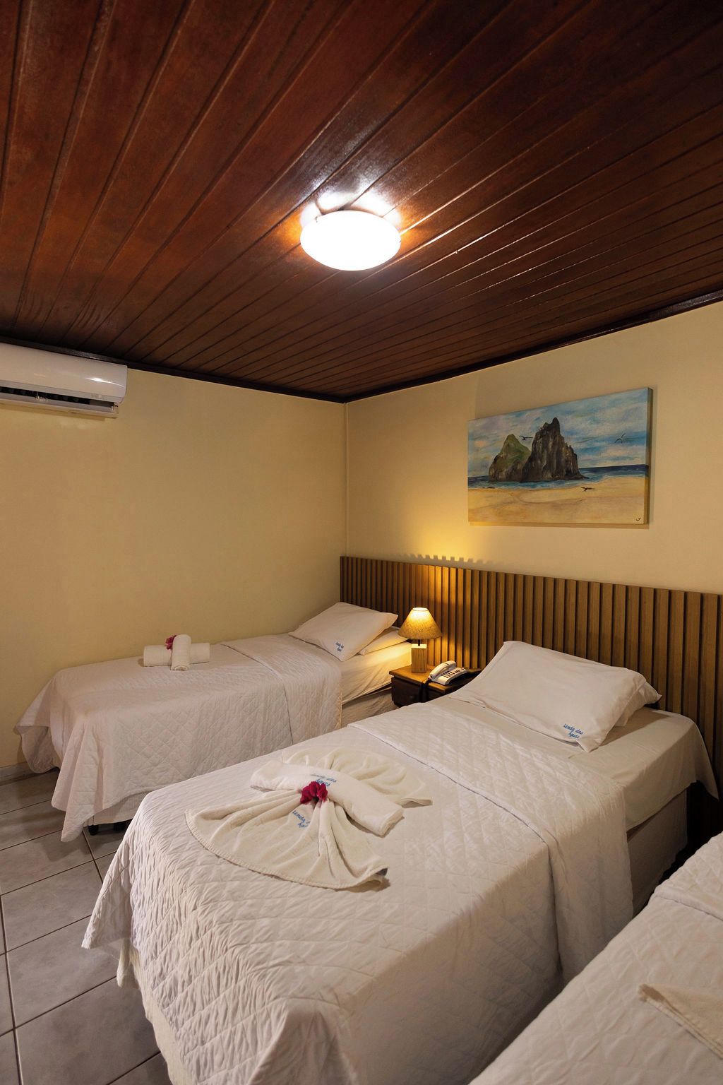 A hotel room with two beds, a painting of a coastal scene, a bedside lamp, and an air conditioning unit.