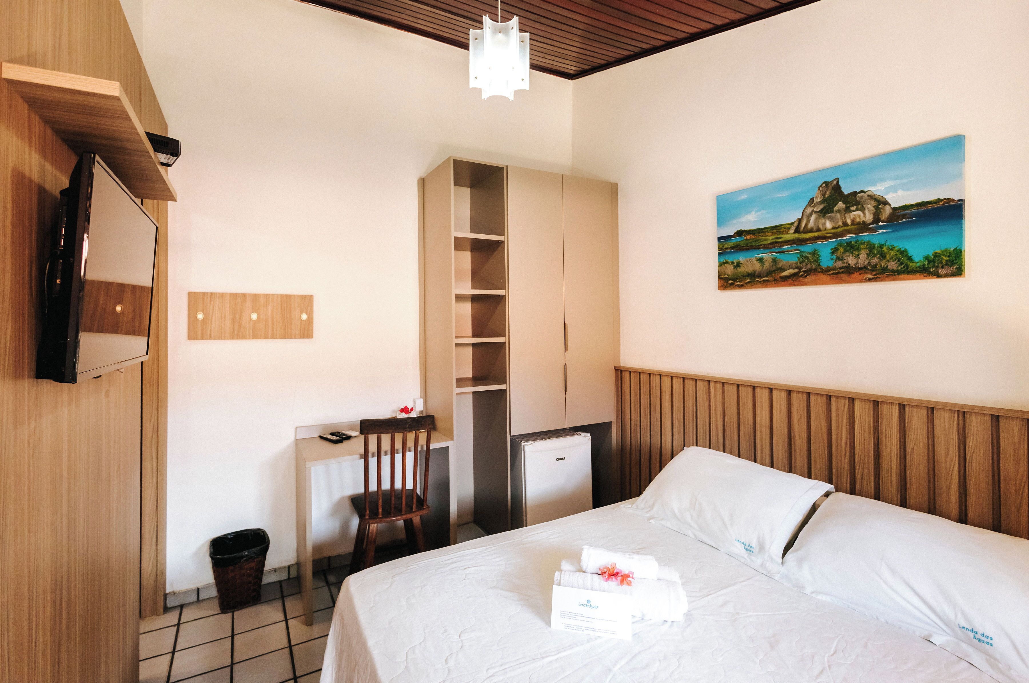 Una habitación de hotel con cama, escritorio, silla, refrigerador pequeño, cesto de basura y un cuadro en la pared.
