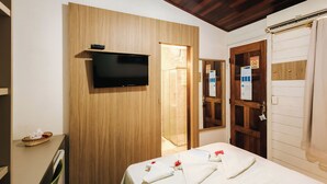 A hotel room with a wooden wall, a flat-screen TV, a wooden door, and a bed with towels and flowers. - Pousada Lenda das Águas (Fernando de Noronha)