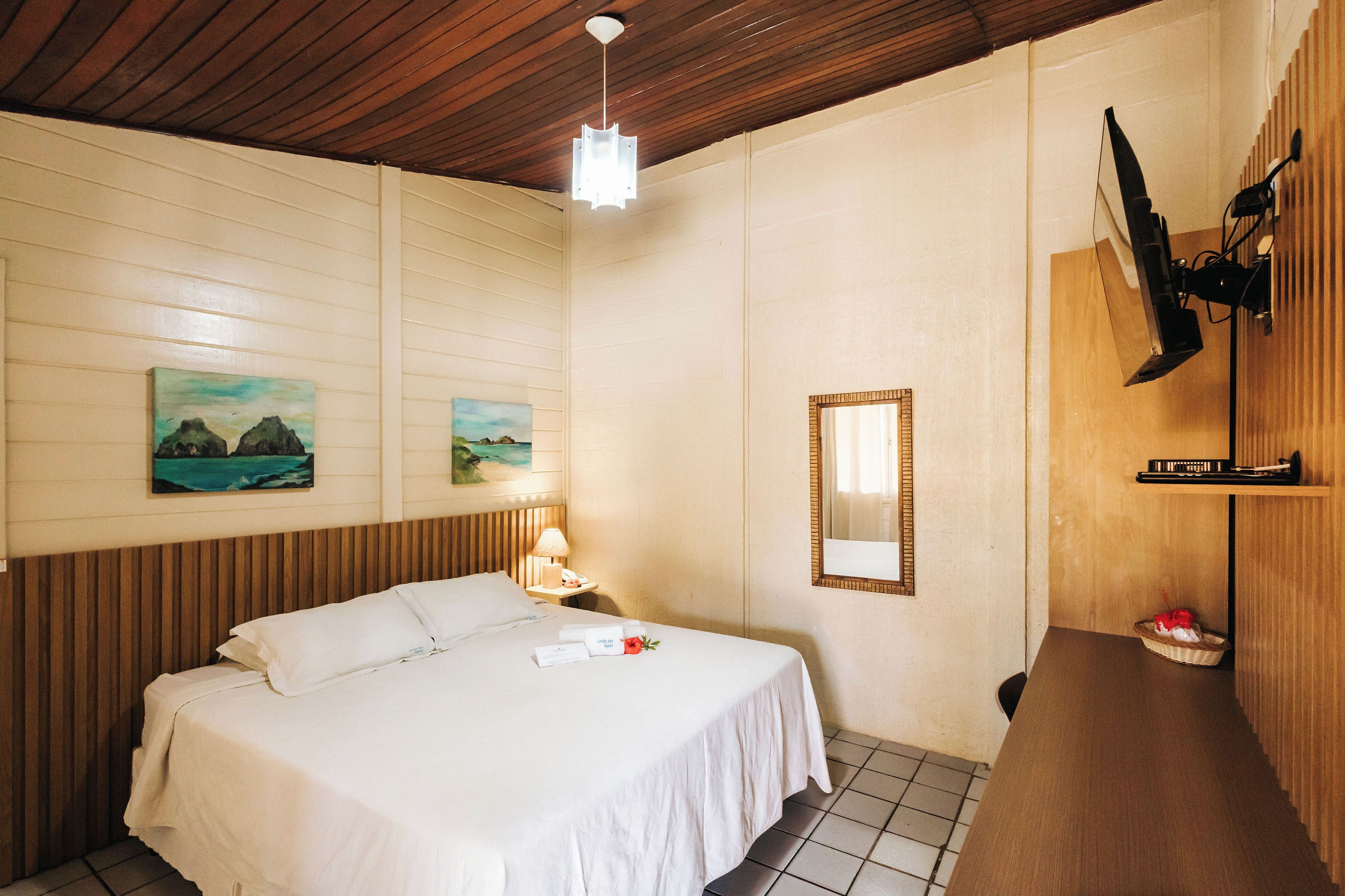 A hotel room with a wooden ceiling, a bed with white linens, a nightstand with a lamp, a wall-mounted TV, and a small shelf with a basket.