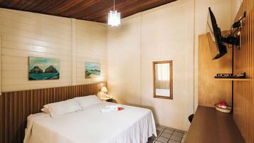 Habitación de hotel con techo de madera, una cama con sábanas blancas, una mesita de noche con lámpara, un televisor montado en la pared y un pequeño estante con una cesta.