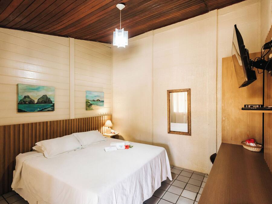 Habitación de hotel con techo de madera, una cama con sábanas blancas, una mesita de noche con lámpara, un televisor montado en la pared y un pequeño estante con una cesta.