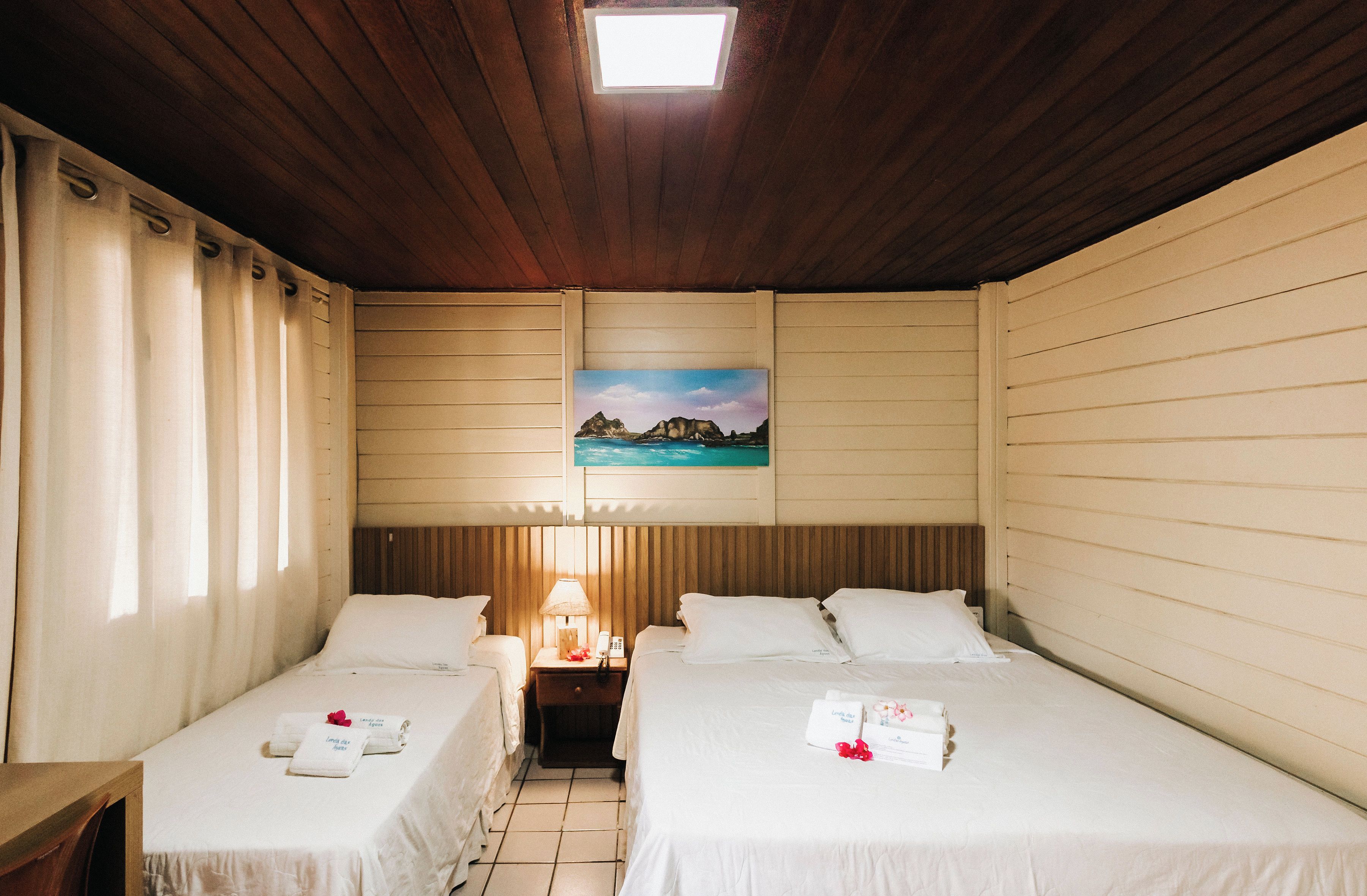A hotel room with two beds, a nightstand, a lamp, and a painting of a coastal scene.