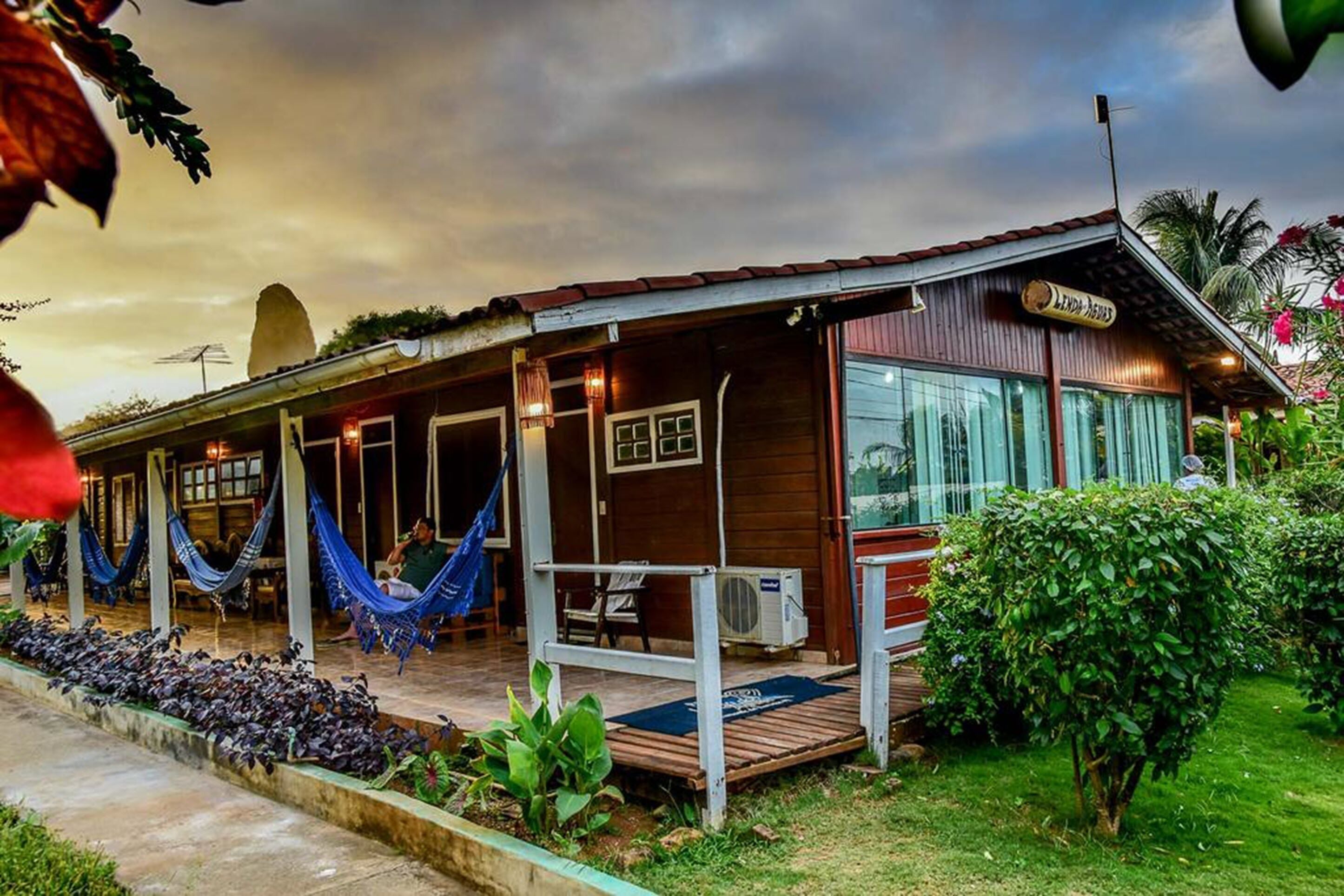 Un bâtiment en bois doté d’un porche couvert, d’une enseigne indiquant « Lindas Agoras », et d’un jardin avec des plantes et un hamac.
