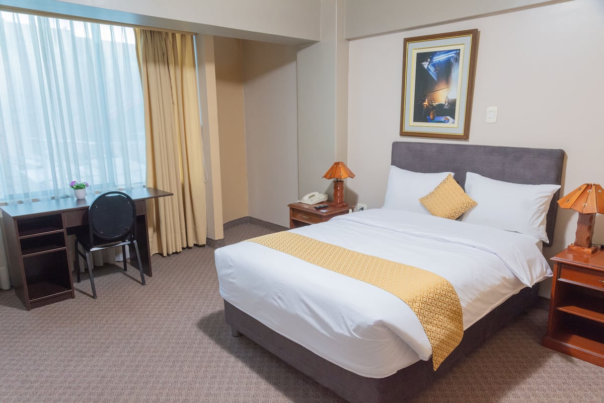 A hotel room with a bed, a desk, a chair, a lamp, and a painting on the wall.
