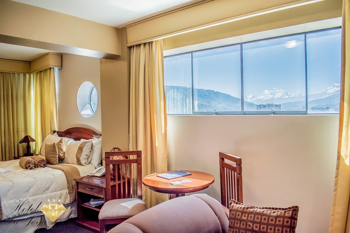 A hotel room with a bed, a round table, two chairs, a small table with a book, a clock on the wall, and a window with a mountain view.
