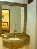 Uma casa de banho com lavatório bancada em granito, um espelho grande e uma cama visível no reflexo.