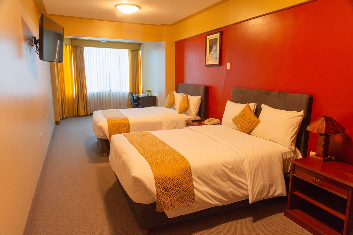 A hotel room with two beds, a TV mounted on the wall, a desk with a chair, and a window with curtains.