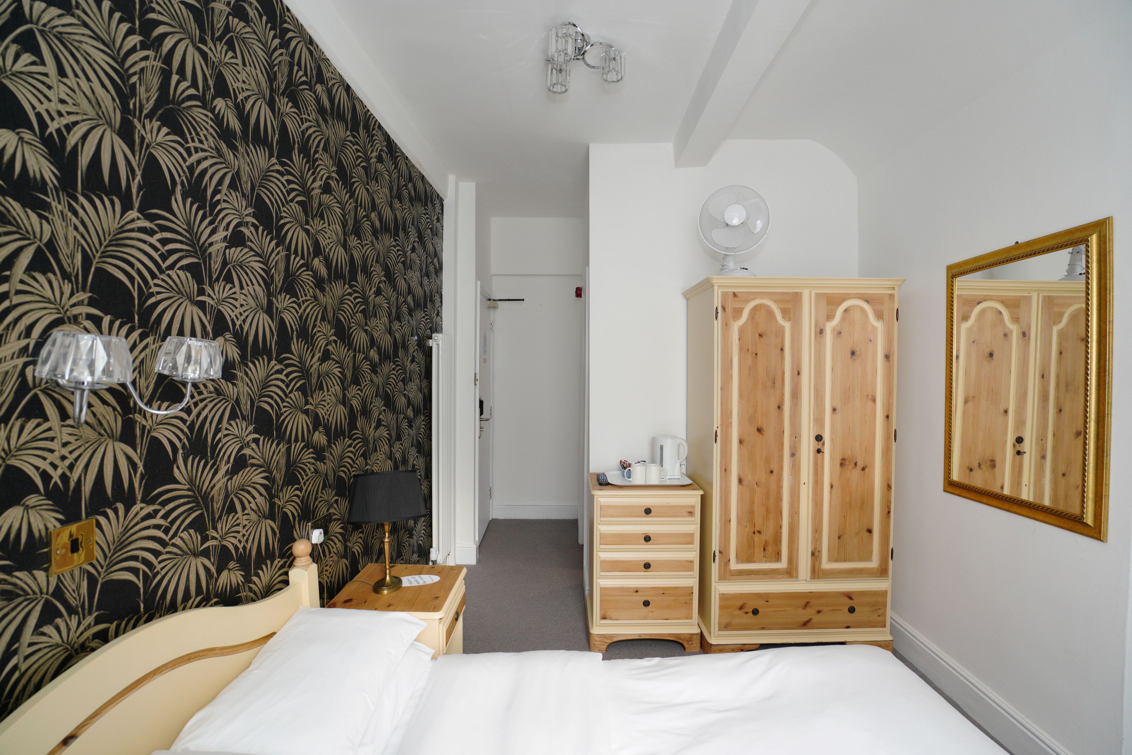 A bedroom with a bed, a wooden wardrobe, a nightstand, a lamp, and a mirror.