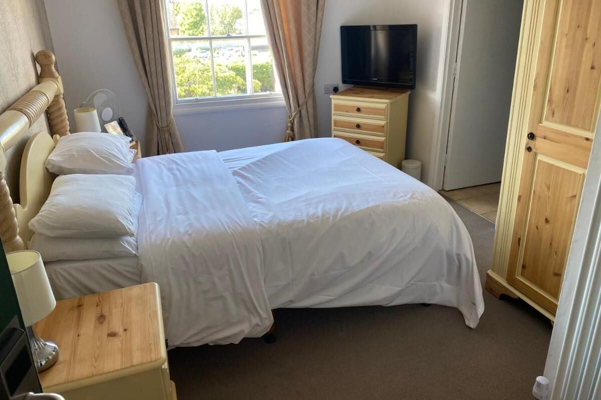A neatly made bed in a room with a window, a television on a wooden stand, and a wooden wardrobe.