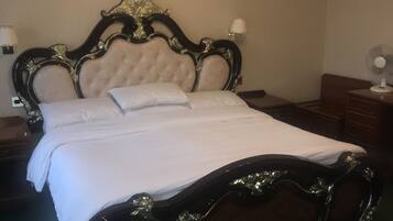 A bed with a dark wooden frame and white bedding, a bedside table with a lamp, and a wall-mounted fan.