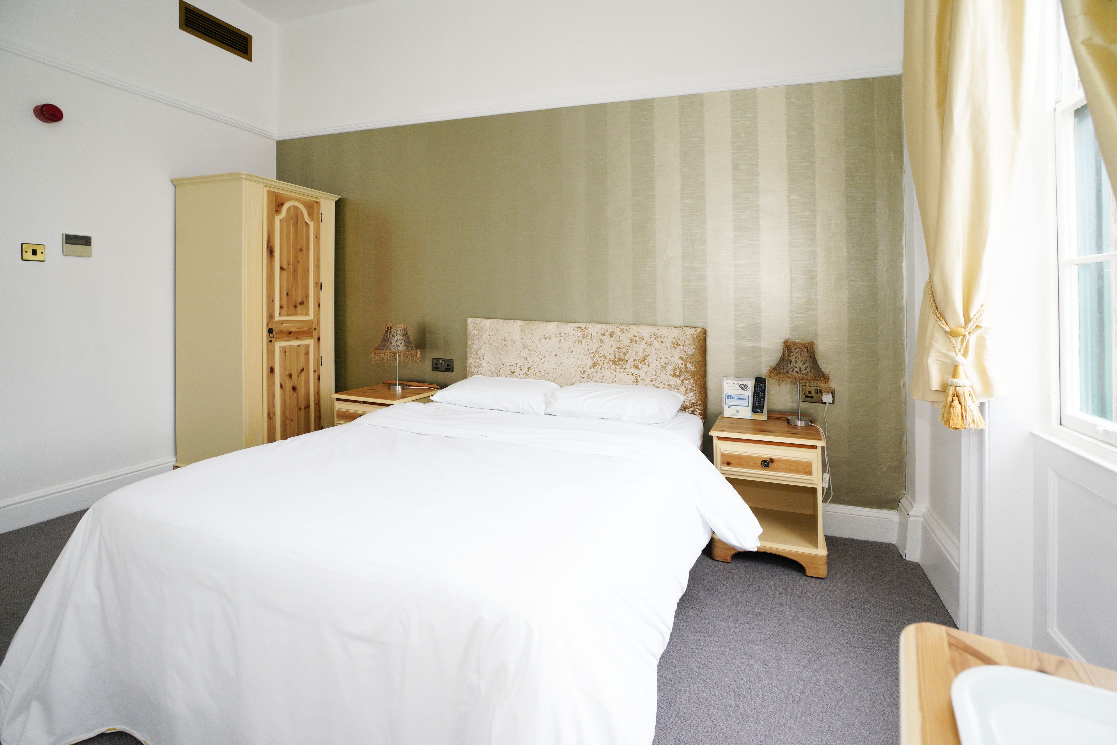A hotel room with a large bed, a wooden wardrobe, a bedside table with a lamp, and a window with curtains.