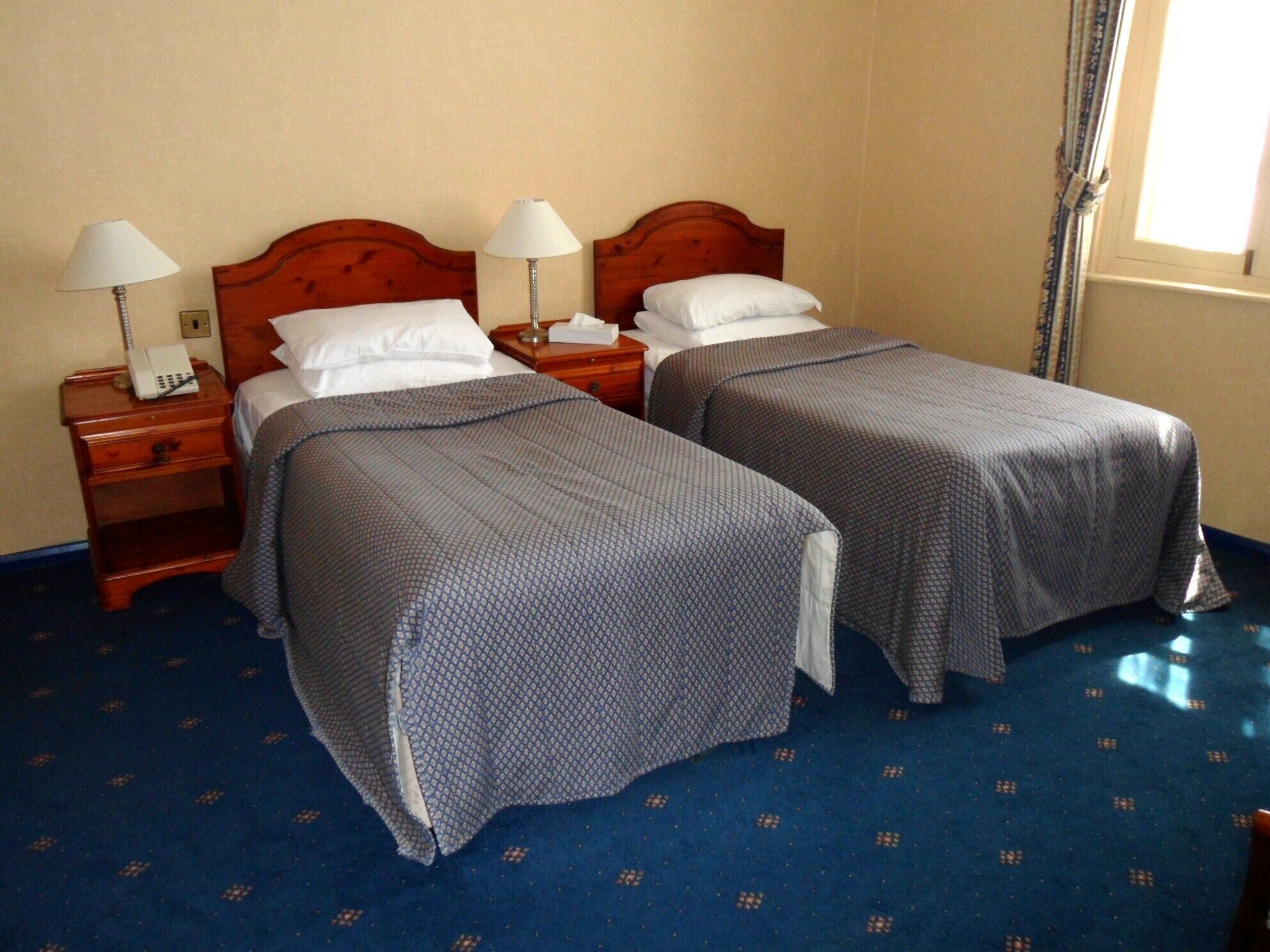 A hotel room with two beds, each with a wooden headboard, white linens, and a blue patterned bedspread. There are bedside tables with lamps and a telephone.